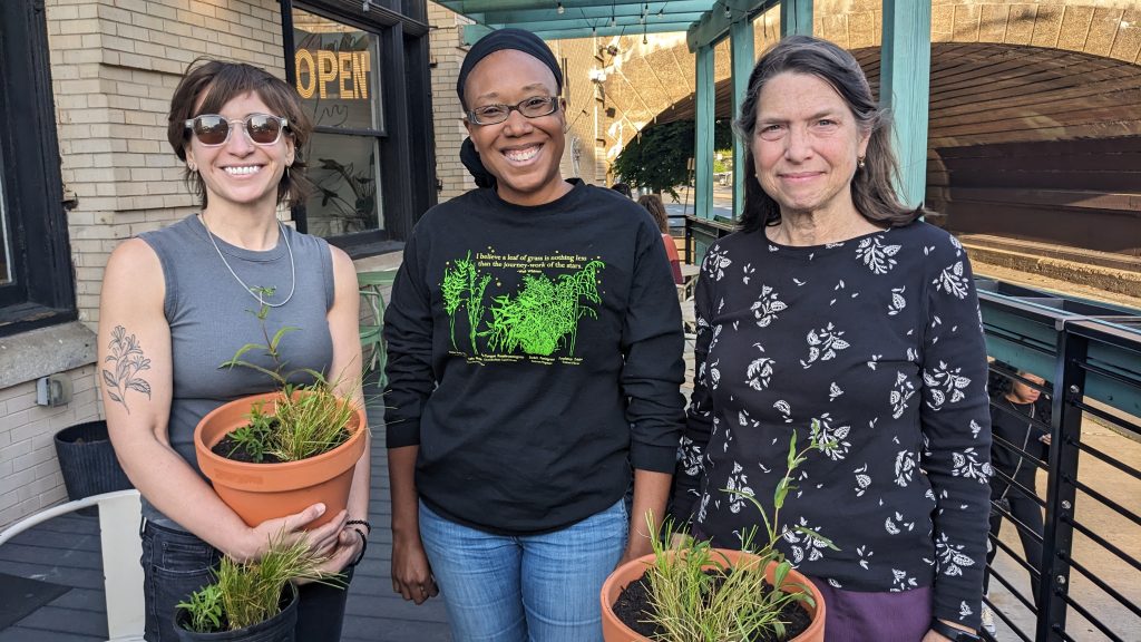 Three women stand outdoors on a patio, each holding potted plants, smiling at the camera in casual clothing.